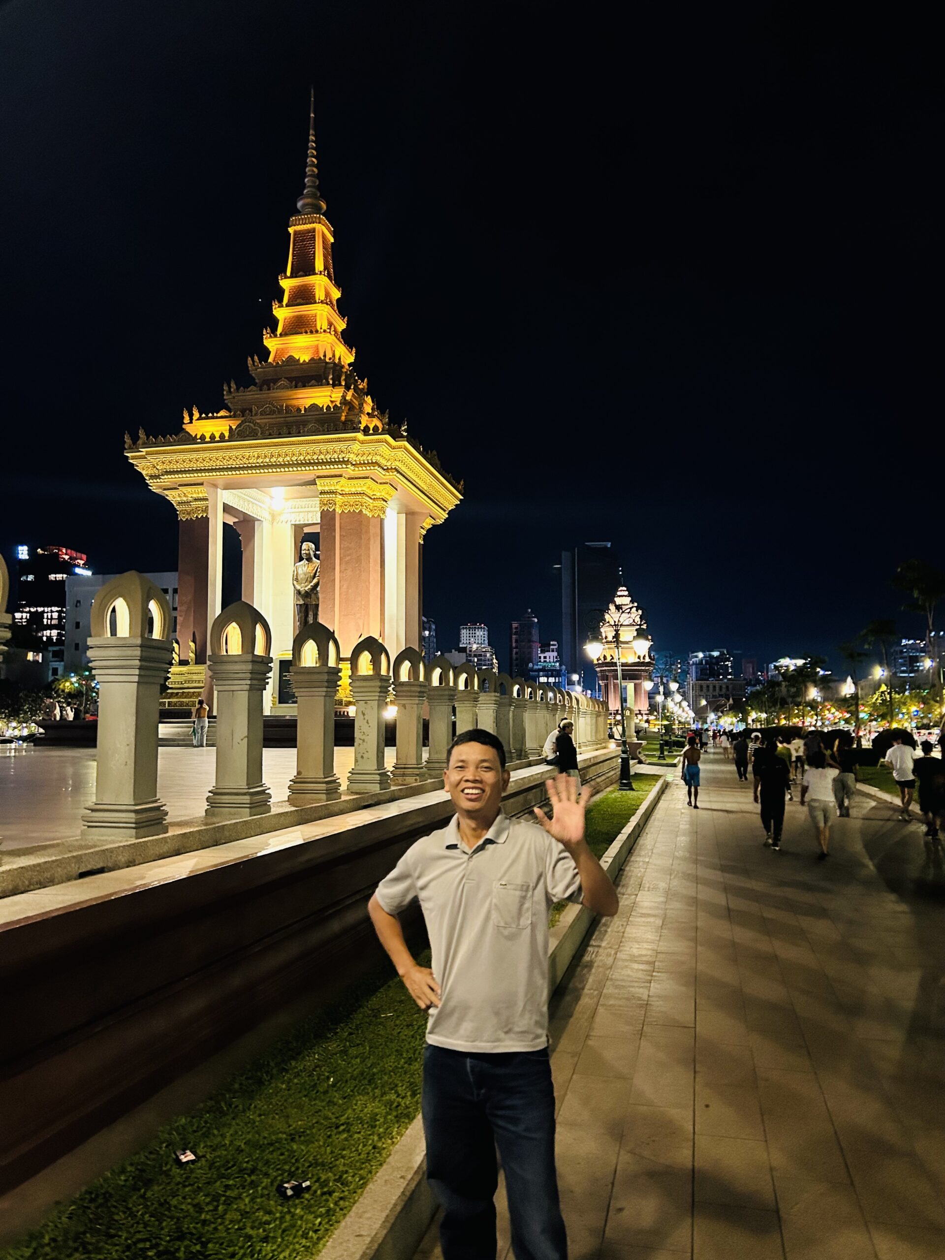 Bunna smiling and waving in front of the Independence Monument in Phnom Penh at night, with the monument beautifully lit and a lively street scene in the background.