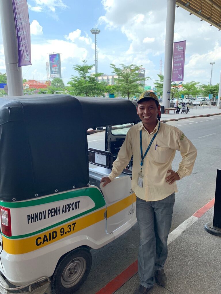 Bunna, a friendly Phnom Penh tuk-tuk driver, smiling with his tuk-tuk at Phnom Penh Airport, ready for safe airport transfers